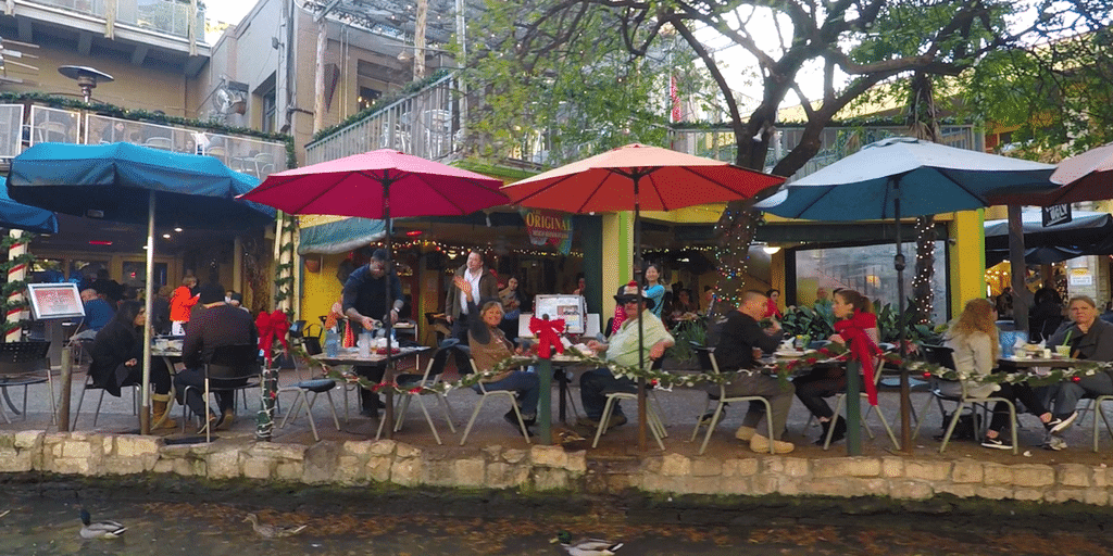 The San Antonio Riverwalk with people eating under umbrellas on the water's edge. 