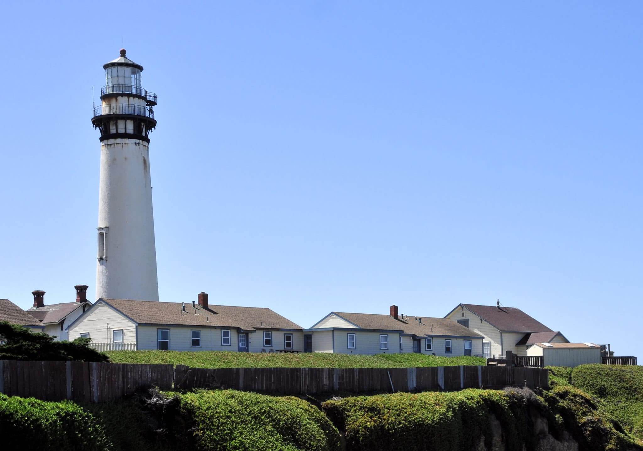 A white building and a lighthouse with clear blue skies in the background and green bushes in the foreground. This is Lighthouse Point, one of the things to do in Santa Cruz. 