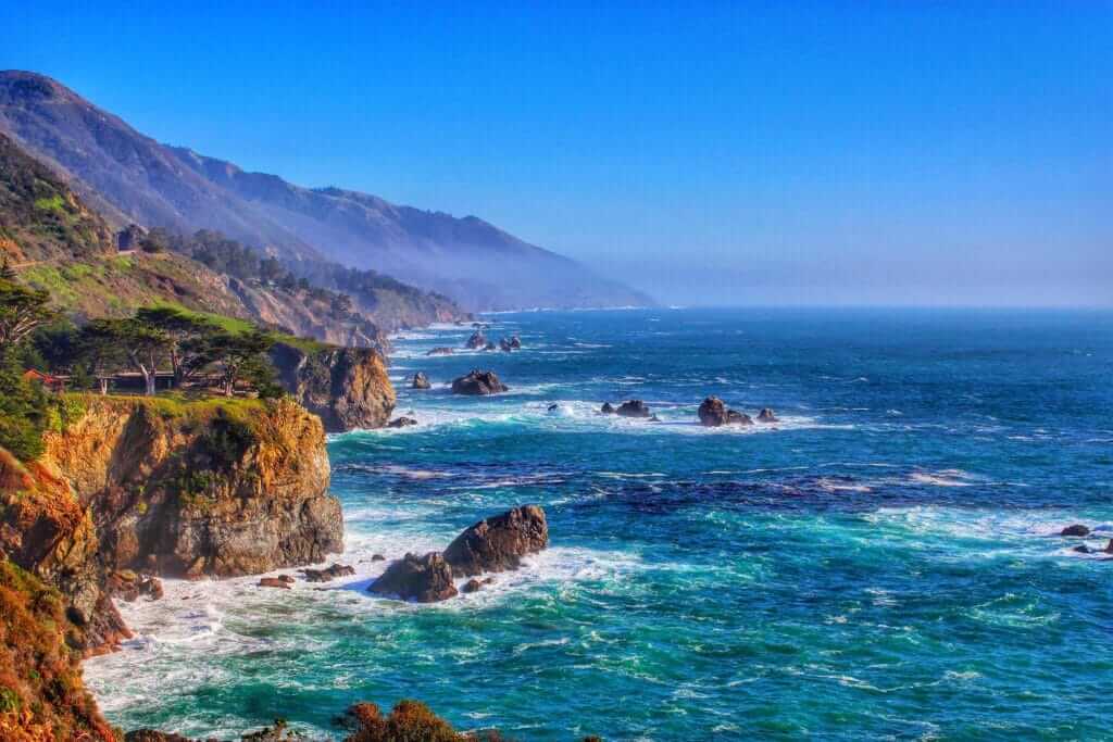 Coastal view of Big Sur with mountains and deep blue ocean crashing against the cliffs, one of the stops on the Pacific Coast Highway Road Trip