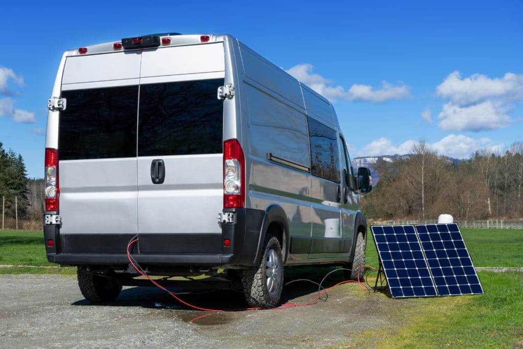 Camper van in an open field with trees and mountains in the background, and with a solar panel plugged in to it. 
