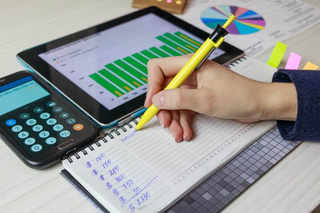 Womans hand holding a pen writing down her budget on paper with a calculator and iPad in front of her. Writing down your spending is the first step to learn ways to save money. 
