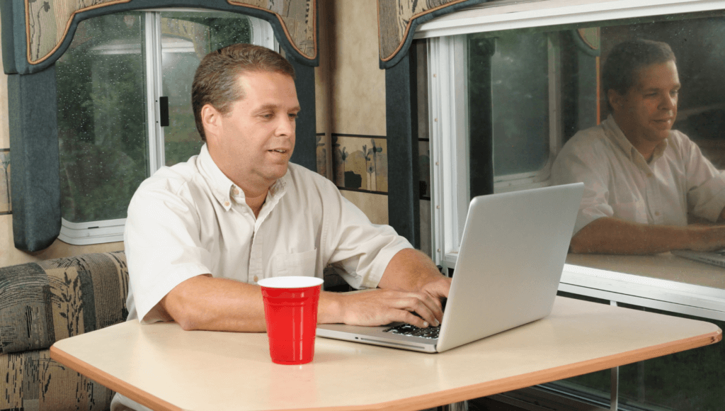 A man sits at the dining table of his RV using a laptop which is connected to an RV hotspot for internet signal.