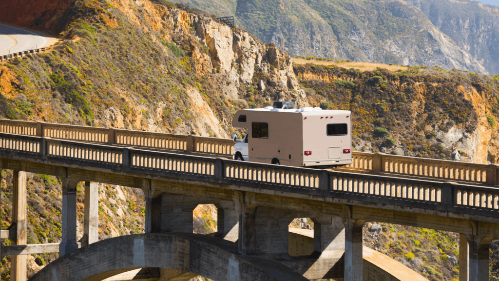 An RV drives across famous Bixby bridge in Big Sur, CA. The cliffs in the background are green and golden with the sunset reflecting off them. 