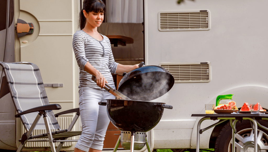 A woman is grilling on a round black grill outside of her camper. She has a chair beside the grill. A table is on the opposite side with cut watermelon and a plate of other fruit.