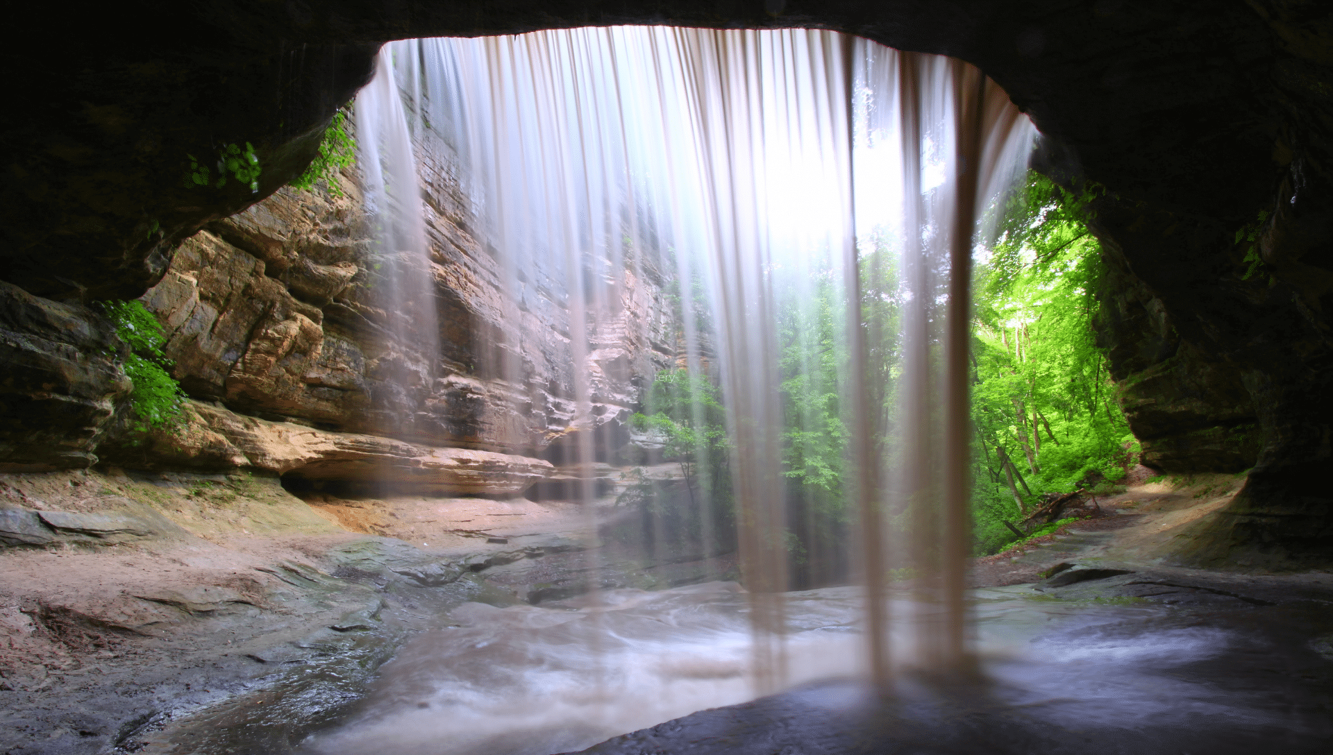 A view from the backside of a waterfall with the water coming down. Sunlight rays and green trees are seen through the water at Starved Rock State Park.