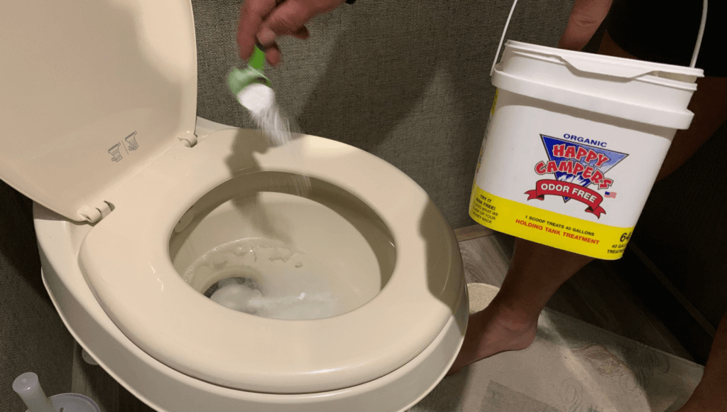 A man stands in an RV bathroom holding a container of Happy Campers RV tank treatment while pouring a scoop of the treatment into the toilet.