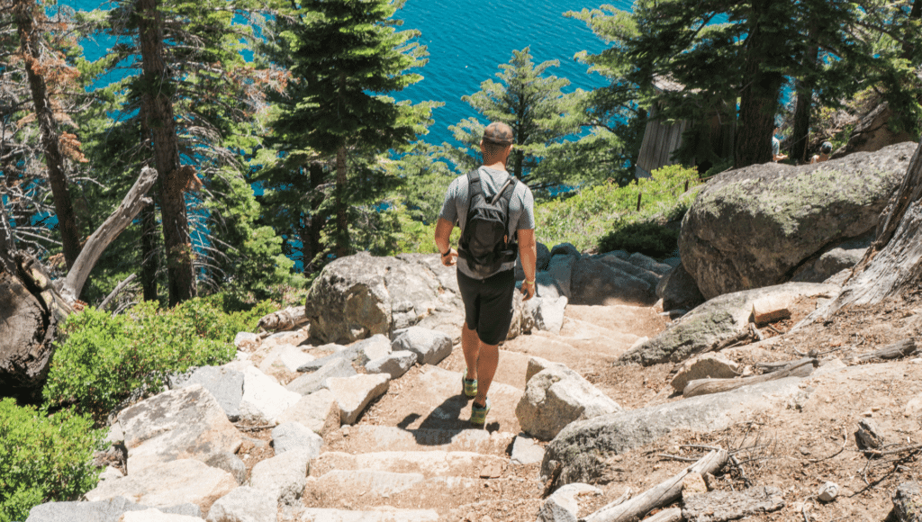A man with a backpack walks down a trail with stone steps amongst rocks while Lake Tahoe camping. Just below him are tall  pine trees and a beautiful view of Lake Tahoe.