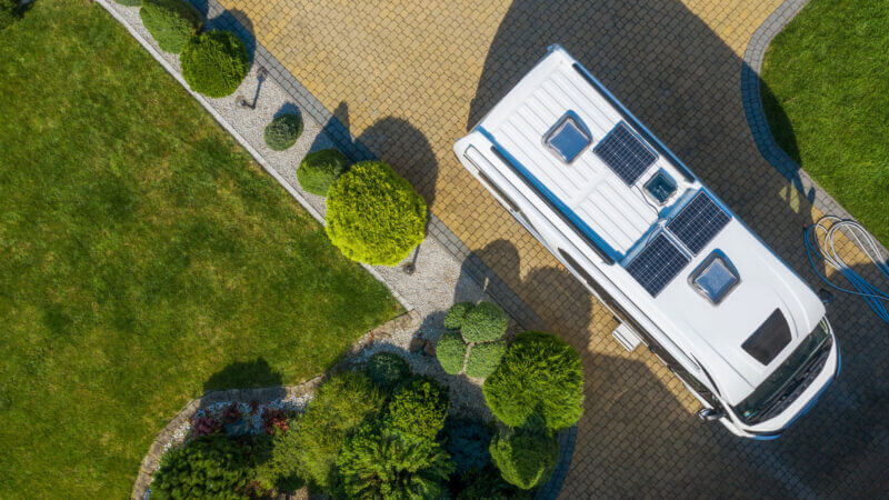 How to know how many solar panels for your RV? An aerial shot of a white RV with 3 solar panels on its roof is parked in a sunny driveway surrounded by grass and small hedges.