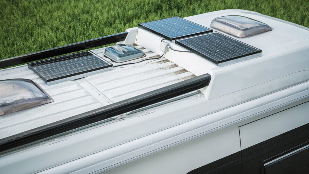 A close up shot of a white RV roof with 3 solar panels installed with grass in the background. 