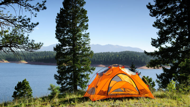 Orange tent camping under some pine trees above a lake with mountains in the background. Ultimate camping hacks to make camping that much better!