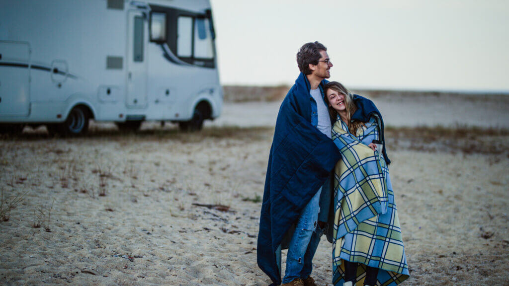 A couple embrace while wrapped in blankets to stay warm outside of their RV which is parked behind them. 