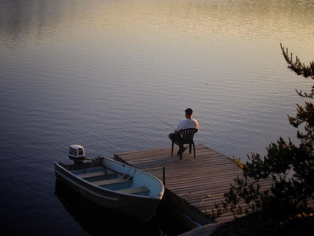 A man sitting in a chair on a dock with a small boat next to him. He has a fishing line in the water. Fishing is a great activity to partake in when Stampede Reservoir camping. 