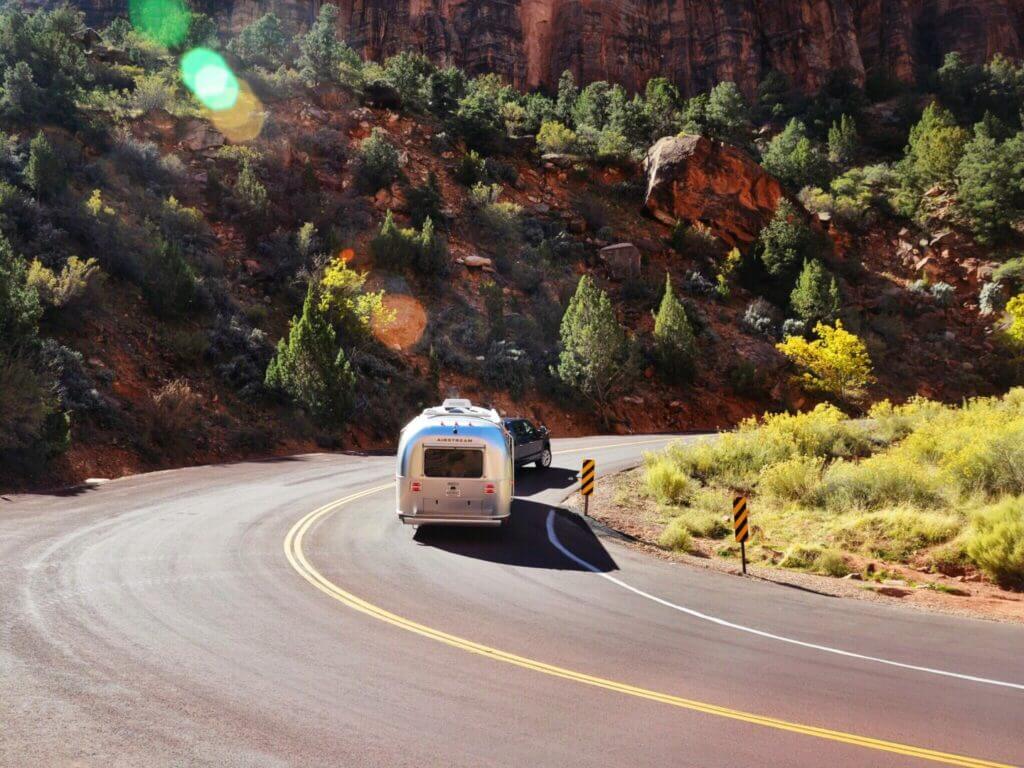 An Airstream brand travel trailer is towed down a winding road lining a rocky cliff.