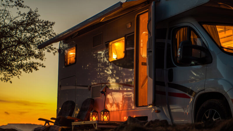 A parked RV with open windows and door parked next to a tree glows in the light orange light from the sunset.