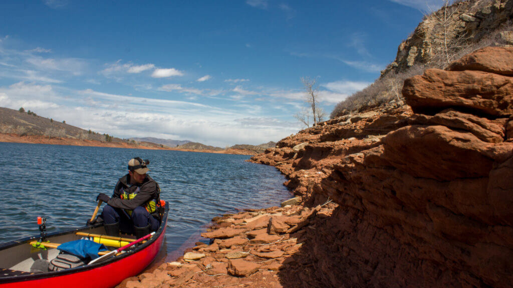 A man rows along Horsetooth Reservoir in a red canoe looking for a place to set up camp along the red rock shore. 