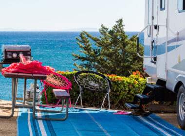 An RV camp is set up at the beach with a picnic table, chairs, and an outdoor mat.