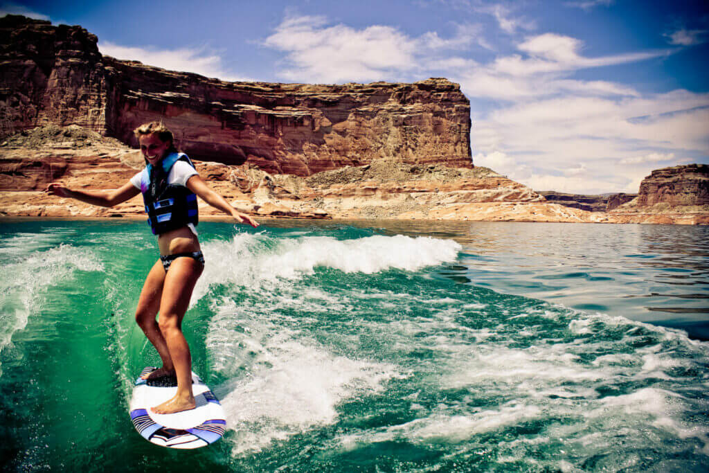 Girl on a board wakeboarding on Lake Powell with tall canyon rocks in the background. This is one of the activities you can partake in when Lake Powell RV camping. 