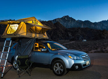 An awesome car camping experience with the tent on the roof of the SUV and it's glowing in the blue light of dusk with mountains and a dark sky fading in the background.