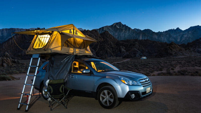 An awesome car camping experience with the tent on the roof of the SUV and it's glowing in the blue light of dusk with mountains and a dark sky fading in the background.