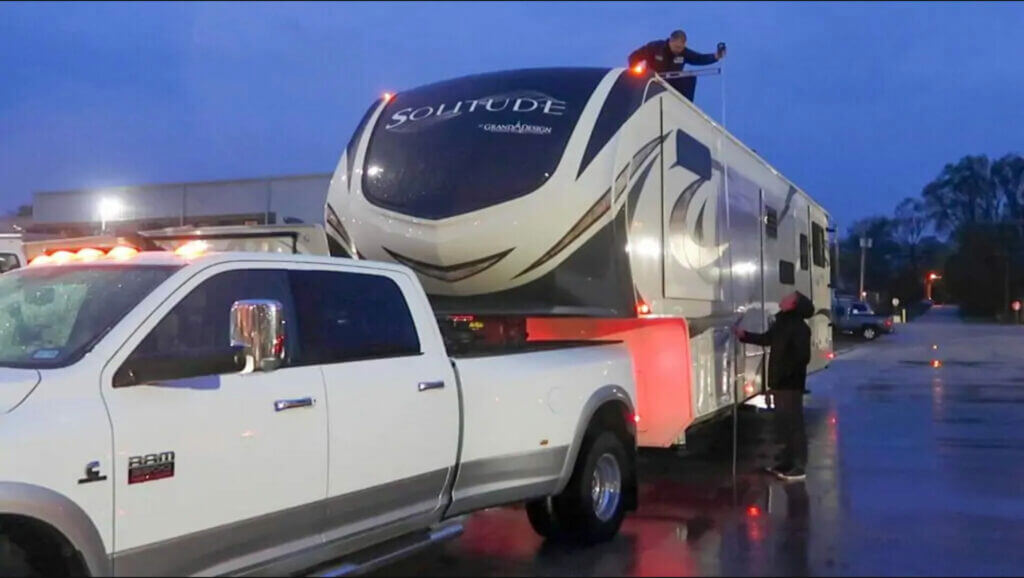 On a dark rainy evening, a white truck is towing an RV and two men are trying to measure the height of the RV with one and the roof and one on the road holding a measuring tape together.