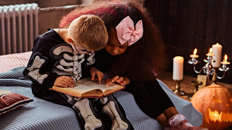 A boy dressed as a skeleton and a girl with a bow in her hair are reading spooky stories for kids next to some spooky halloween candles.