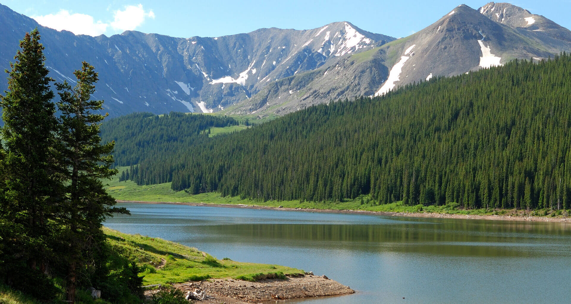 Green Mountain Reservoir on a blue sky day with mountains in the background, blue water, and green pine trees in the forefront. Green Mountain Reservoir is a hidden camping gem in Colorado.
