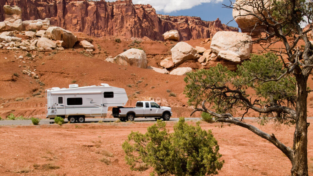 In a red rocky desert a white truck tows a small white travel trailer that may not have a bathroom installed in it. 