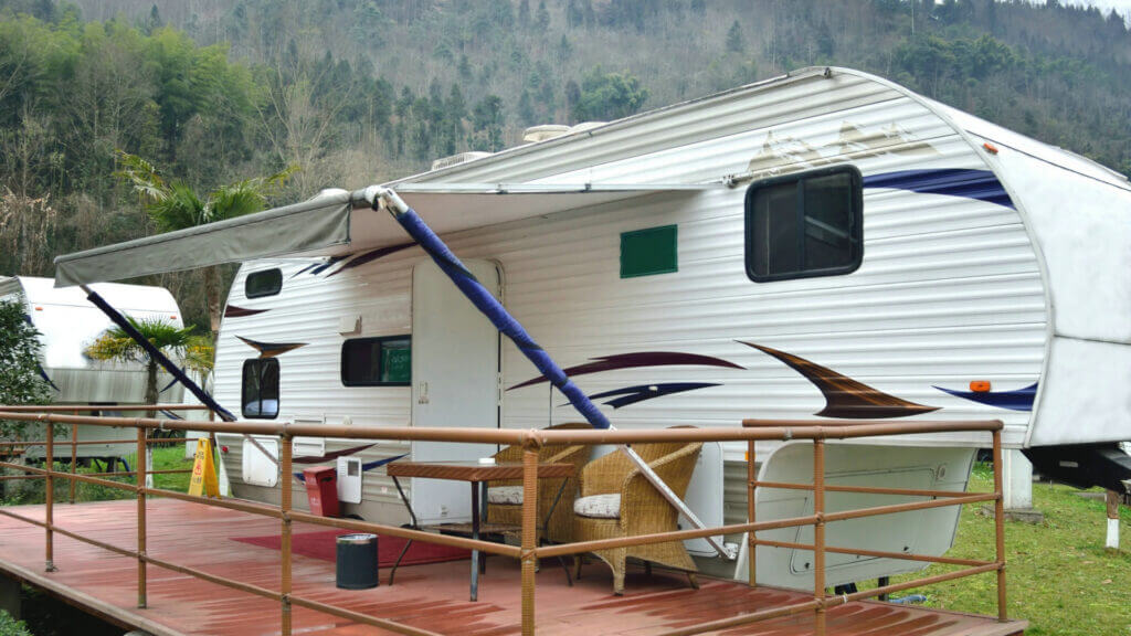 A white RV is parked against the mountains with a deck built up against it because the RV is being used to live in permanently. 