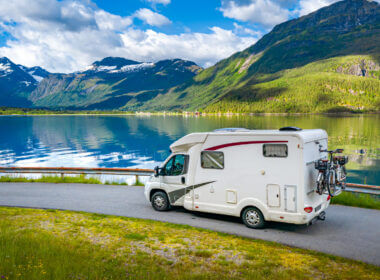 An RV drives along a roadway set against a lake and green and blue mountains.