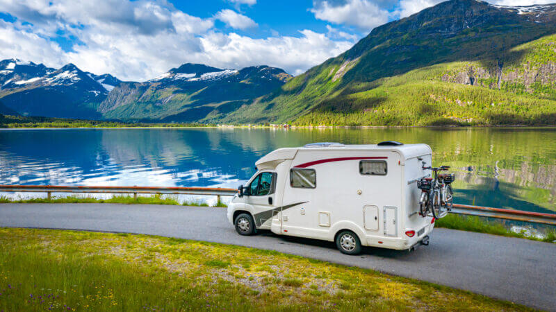 An RV drives along a roadway set against a lake and green and blue mountains.