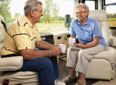 An older couple sit comfortably in their RV recliners and relax while drinking coffee.