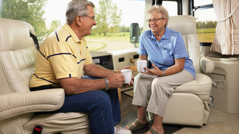 An older couple sit comfortably in their RV recliners and relax while drinking coffee.