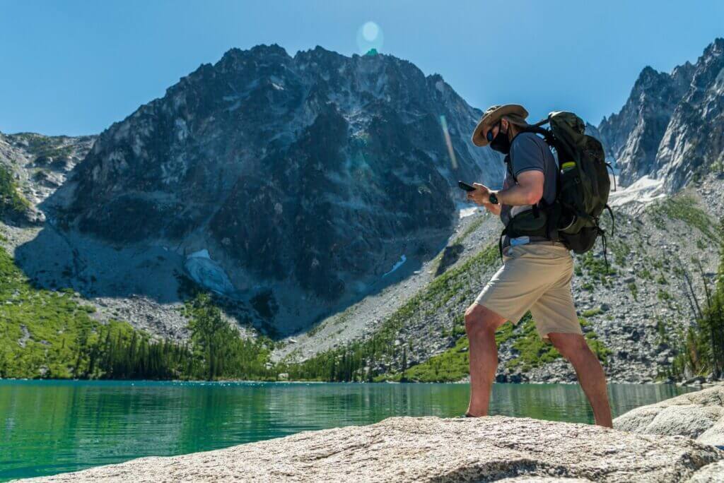 Man standing in front of a beautiful lake with a mask on looking down at his phone