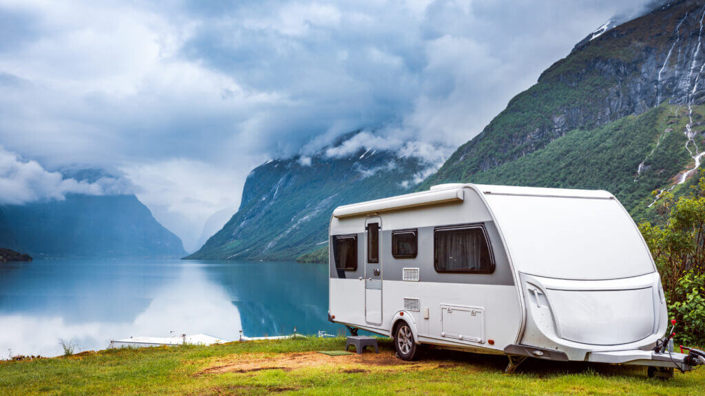 A trailer is left unattended next to a stunning lake and mountains with waterfalls streaming down the sides, and it is safe from thieves because it is secured with a trailer hitch lock.