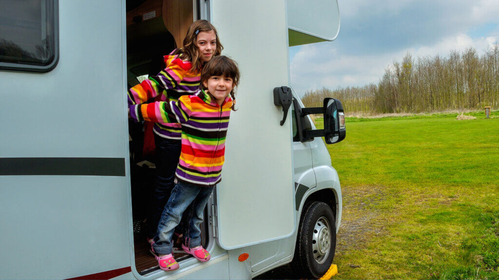 Two girls lean out of the doorway of an RV that is parked in a grassy field. It is parked on a yellow leveler block to help with the uneven terrain. Which is better: Camco vs Lynx levelers?