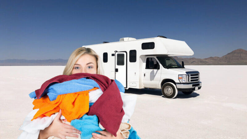 A woman holds a stack of laundry in front of an RV parked in the desert. She can use one of the best portable washing machines anywhere she parks in her tiny space!