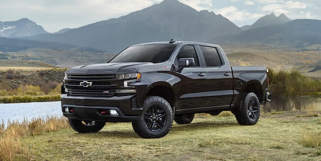 A black Chevy Silverado 1500 is parked on a grassy area near a river among the mountains. 