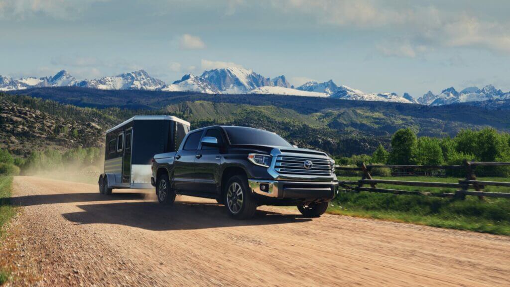 A Toyota Tundra tows a small horse trailer across a dirt country road with snow capped mountains loom in the background. 