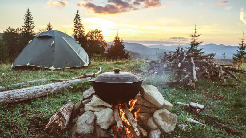 A scenic mountain camping scene with a cast iron pot on top of a fire and the tent among the trees as the sun rise. Try these camping tips and tricks to solve common camping problems!