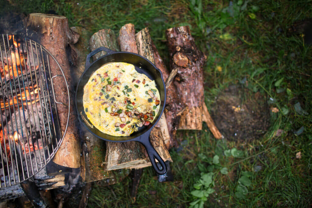 A campfire breakfast with eggs and peppers in a cast iron skillet sitting next to a fire. 