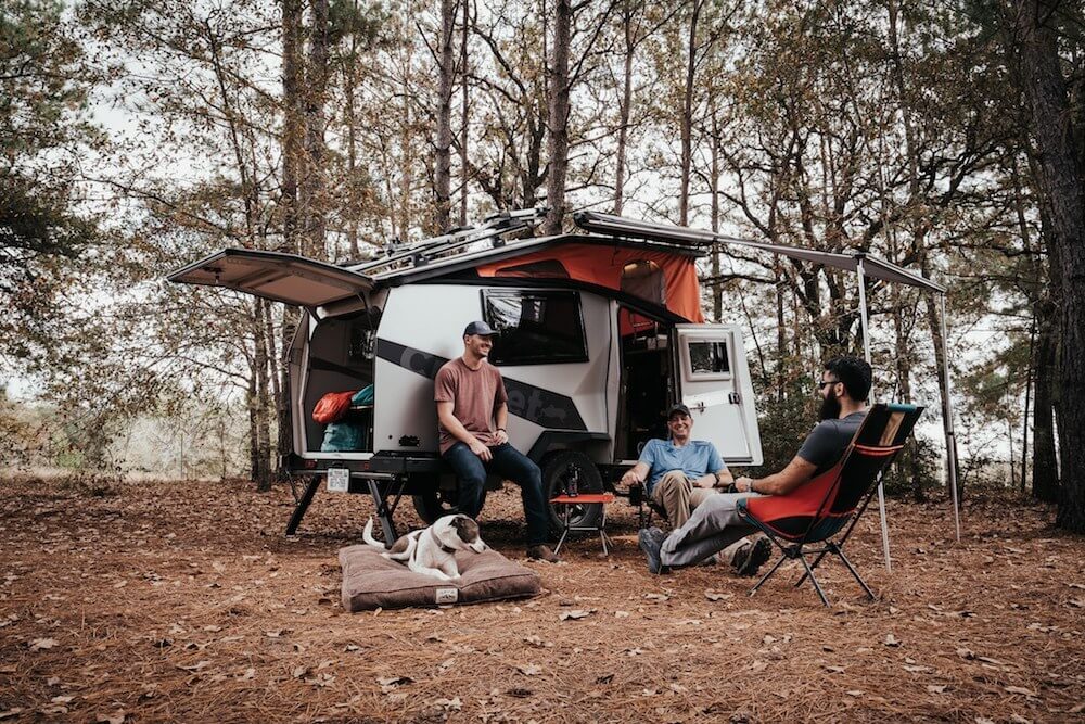 Three men and a dog sitting outside in the woods with their off-road trailer in the background