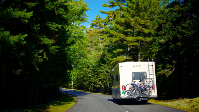 An RV with a bumper bike rack drives along a highway in a green forest.