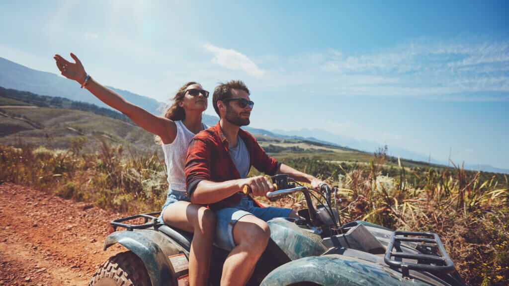 A man and woman happily ride an ATV on a clay road in the sun. A toy hauler is an RV that allows you to travel with your ATVs or motorcycles or other 'toys'.