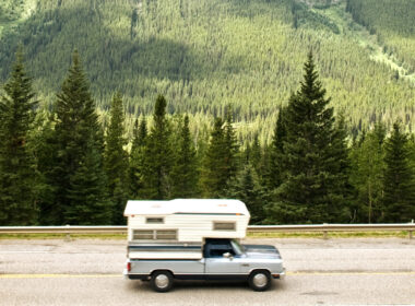 A truck camper cruised along a highway next to green mountain forests.