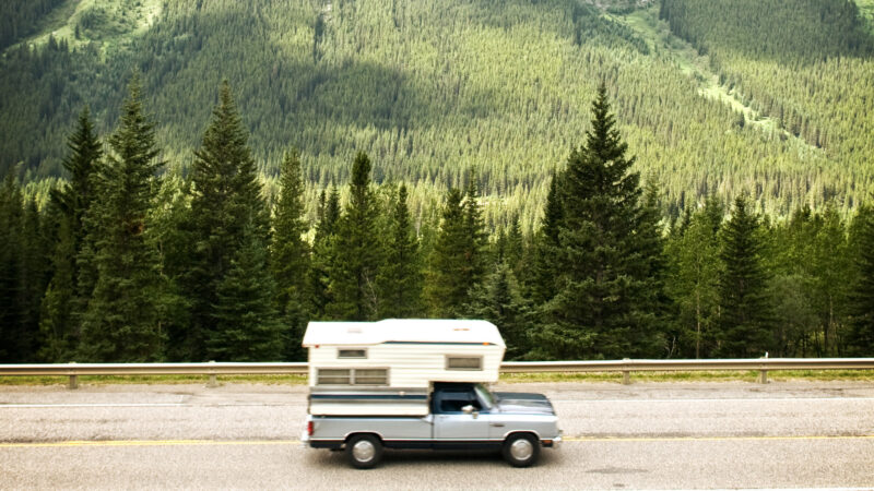 A truck camper cruised along a highway next to green mountain forests.