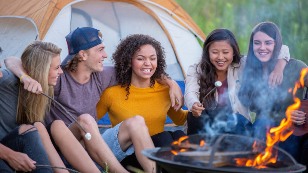 A group of adults laugh as they play campfire games and roast smores outside.