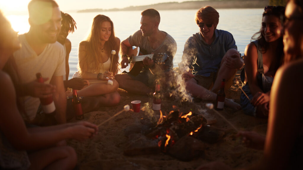A group of young adults enjoy smores, music, and drinking games around a campfire on a lake shore at sunset.