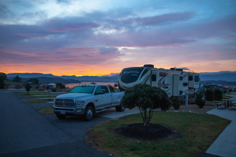 A white dodge truck and Solitude RV sit parked in a campground with a beautiful sunset in the background.