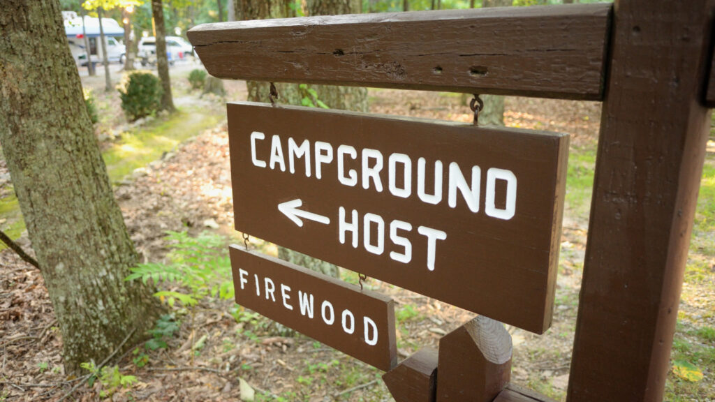 A campground host sign in a campground with RVs in the background. Hosting for just a place to stay and no additional income is a common workamping regret. 