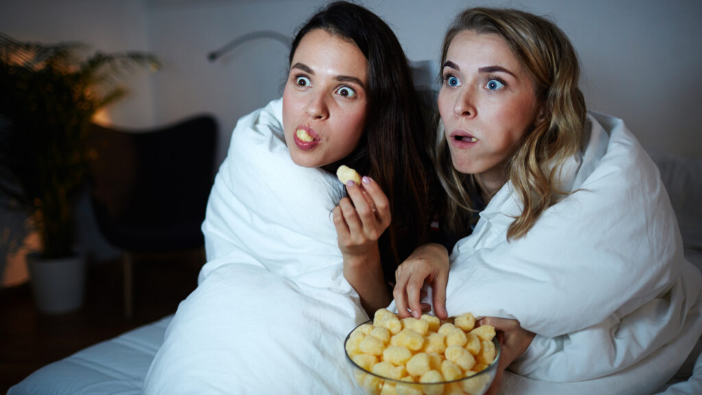 Two girls share a blanket and puff snacks and look shocked while watching a movie.